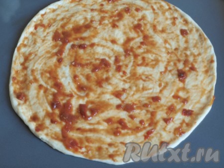 Разделить готовое тесто на две одинаковых части. Одну часть убрать в пакет, чтобы тесто не заветривалось. Можно тесто убрать в холодильник, пару дней оно может полежать там. Если не собираетесь сразу готовить 2 пиццы, то можно убрать тесто в морозилку и воспользоваться им в другое время. Противень смазать растительным маслом. Из оставшейся части теста раскатать на противне достаточно тонкую круглую лепёшку. Тесто прекрасно формируется руками. Затем лепёшку нужно смазать оливковым маслом, а сверху - томатным соусом.
