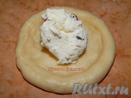 Выложить тесто на стол. От теста отрезать кусочки весом, примерно, по 80-100 грамм. Получившиеся кусочки теста скатать в шарики. С помощью скалки придать шарикам из теста круглую форму, в центре сделать углубление. Получившиеся заготовки для ватрушек аккуратно выложить на противень, застеленный фольгой (или пергаментом). В центр каждой ватрушки выложить по 1 столовой ложке начинки из творога и изюма.