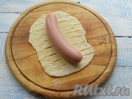 На столе, присыпанном мукой, каждую часть теста раскатать достаточно тонко в пласт овальной или прямоугольной формы по длине сосиски. Сосиску очистить и поместить по середине теста. По бокам сделать по 6-8 разрезов.