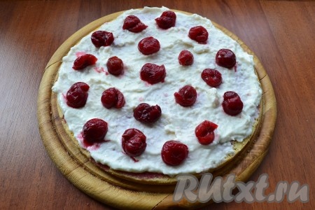 Далее собрать бисквитный торт, для этого смазать кремом нижнюю часть бисквита, выложить половину ягод вишни из начинки. 