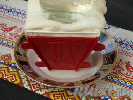 Поставить пасочницу в тарелку для стекания сыворотки, края подвернуть наверх. Сверху поставить тарелку и на неё груз весом 500 грамм. Всё вместе поставить в холодильник на 12-15 часов. За это время слить сыворотку 2-3 раза.
