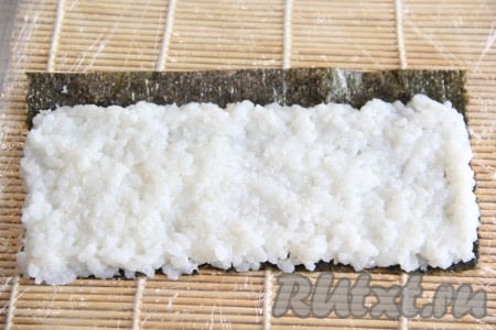 Каждый лист нори разрезать на 2 части. На подготовленную циновку поместить лист нори блестящей стороной вниз. На нори выложить немного риса и разровнять его по всей поверхности,&nbsp;оставляя свободный край листа нори (примерно 1 сантиметр).