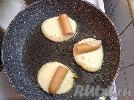 На каждый кружок теста положить кусочек сосиски немного ближе к одному краю. Тем временем оладушки начнут поджариваться с одной стороны.