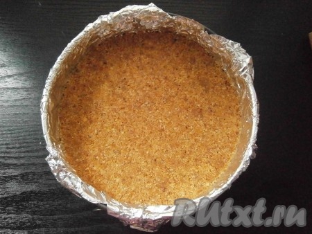 Разъёмную форму диаметром 18-19 сантиметров застелить пищевой плёнкой (или фольгой), чтобы концы свисали с краёв формы. Выложить крошку с маслом и хорошо её утрамбовать по дну. Поместить форму в морозилку на 30 минут.