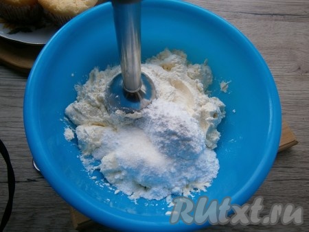 Дать капкейкам полностью остыть, а пока приготовить творожный крем. Для этого к мягкому творогу добавить сметану, перемешать. Всыпать сахарную пудру и ванильный сахар, взбить массу до однородного состояния погружным блендером.
