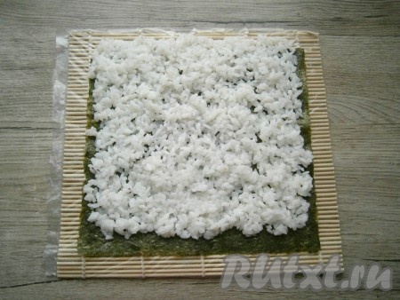 Лист нори расположить на циновке (лучше циновку обернуть пленкой) шершавой стороной кверху. Рис выложить на нори тонким слоем так, чтобы снизу он не доходил до края листа на 1 см, а сверху - наоборот, заходил на 1 см за край листа нори. Это нужно для того, чтобы после сворачивания рис хорошо закрепился и не было голых участков нори.
