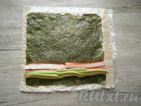 На противоположный край листа нори, отступив от края, примерно, 1 см выложить полоску из авокадо и полоску из нарезанных крабовых палочек.
