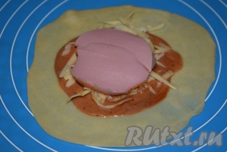 На один раскатанный кружок выкладываем 1 чайную ложку приготовленного соуса. Размазываем соус по поверхности теста, оставляя края чистыми. На соус насыпаем часть сыра. Поверх сыра выкладываем кружочек колбасы.
