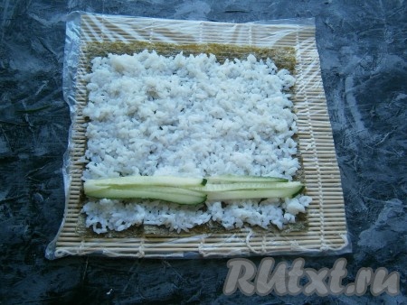 Равномерно влажными руками распределить рис по всему листу, не доходя до верхнего края нори около 0,5-1 см. Огурец нарезать длинными и тонкими полосами, выложить его на рис ближе к нижнему краю (как на фото).
