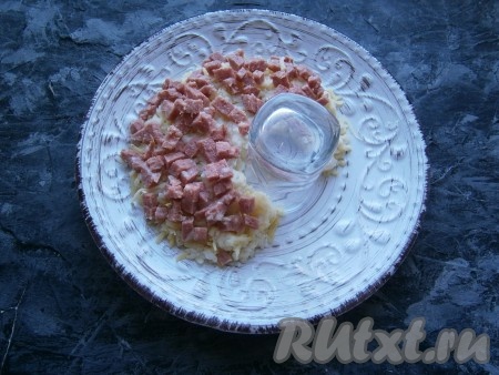 На часть плоской тарелки (ближе к краю) поместить рюмку. Вокруг рюмки выложить очищенный и натёртый на средней (или мелкой) тёрке варёный картофель в виде полукруга, посолить его, поперчить, смазать майонезом. На картофель выложить нарезанную мелкими кубиками колбасу.
