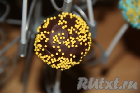 Обмакнуть каждый шарик в шоколад (тёмный или белый) и обвалять в посыпке. Поставить пирожные на палочке на подставку (или в стакан), дождаться, чтобы шоколад схватился, а затем отправить шоколадные кейк-попсы в прохладное место.
