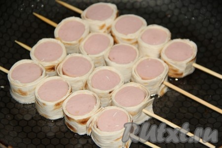 В сковороду влить немного растительного масла и выложить сосиски в лаваше на шпажках.
