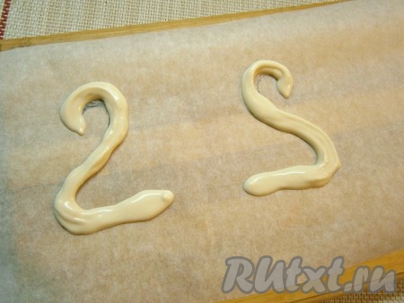 Теперь можно приступить к украшению тортика. Для того чтобы приготовить шеи с мордочками лебедей, нужно растопить на водяной бане белый шоколад, перелить его в одноразовый кондитерский мешок, обрезать кончик. На лист пергамента, размещённый на ровной досочке, выдавить шоколад в виде кривой, похожей на цифру "2", и также выдавить ещё одну кривую в виде "2" как бы в перевёрнутом виде (как на фото). Лучше предварительно нарисовать на пергаменте рисунки - так легче наносить шоколад. Отправить доску с пергаментом в холодильник до полного застывания шоколада.
