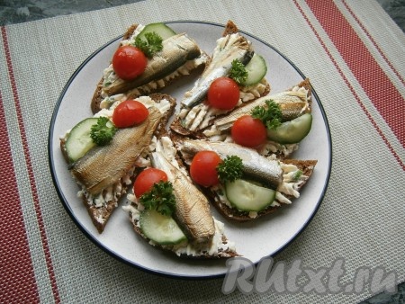 Поверх сырной намазки, ближе к одному из краёв бутерброда, выложить 1 шпротинку (на каждый бутерброд нужно выкладывать по 1 рыбке). Далее украсить бутерброды половинками помидоров черри (или кусочками обычного помидора) и полукружочками свежего огурца, а также зеленью петрушки.
