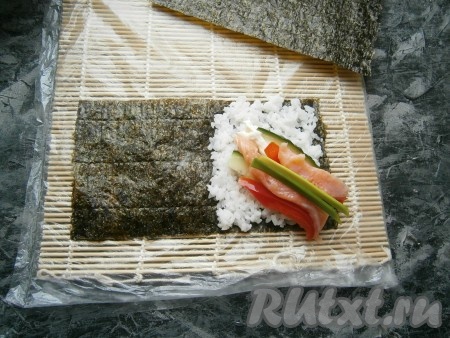 Выложить по 2-3 кусочка нарезанных рыбы, перца, огурца и перьев зеленого лука, как показано на фото. Завернуть этот лист нори в виде кулёчка, начиная с верхнего правого уголка.
