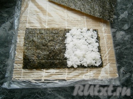 Каждый лист нори разрезать на 2 части. Взять половину листа нори, выложить шершавой стороной вверх, затем равномерно заполнить половину поверхности 40 граммами риса (как на фото).
