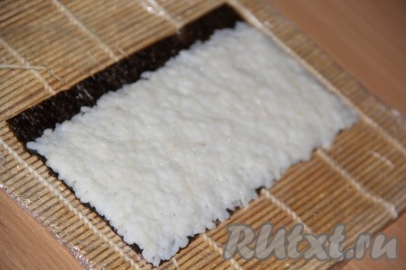 Разрезать лист нори на две части. Половину листа нори выложить на циновку блестящей стороной вниз. Равномерно распределить рис по листу, оставив с одного края 1 см листа свободным. Толщина риса должна быть, примерно, 5 мм.

