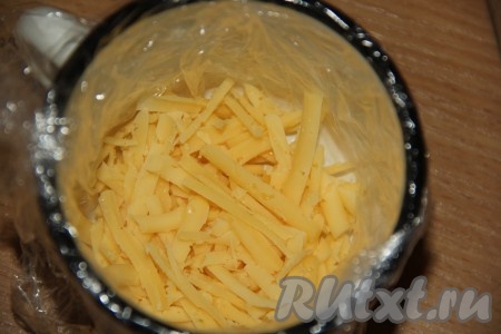 Кружку выстелить пищевой плёнкой так, чтобы концы плёнки свисали с краёв (можно сделать бортики из любой жестяной банки или использовать небольшую форму для выпечки кулича). Выложить на дно кружки половину сыра, натёртого на крупной тёрке.