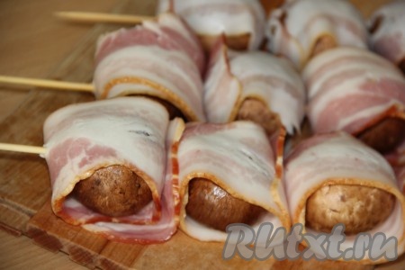 Грибочки слегка посолить. Полоску бекона разрезать на 2 части. Обернуть шампиньоны в бекон и нанизать на деревянные шпажки. Шпажки нужно предварительно замочить на 30 минут в воде. Вместо шпажек можно использовать шампуры.