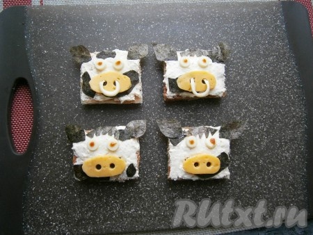 Теперь осталось бутерброды украсить в виде мордочек быков. Для этого из листа нори вырезать ушки, чубы и хаотичные кусочки в виде пятен, оформить на бутербродах. Нос вырезать из тонких ломтиков твёрдого сыра, в которых сделать отверстия (ноздри) колпачком шариковой ручки, выложить на бутерброды. Глазки также колпачком (или чем-то другим) выдавить из белого сыра, разместить на бутербродах, капнуть точки кетчупом, имитируя зрачки. И, по желанию, из белого сыра вырезать тонкие полосочки и вставить их в ноздри в виде колец.
