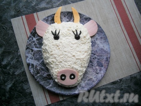 Осталось салатик украсить, для этого из кружочков колбасы вырезать нос и уши быка, разместить на блюде. Из оливок сделать глаза и ресницы, а также ноздри, которые вставить в отверстия, вырезанные в кружочке колбасы, который имитирует нос, выложить на салат. Из сыра вырезать рога и чёлку быку.
