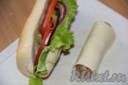 Булочку для хот дога разрезать вдоль. Вложить в разрез лист салата, тонкие ломтики помидора и кольца лука.
