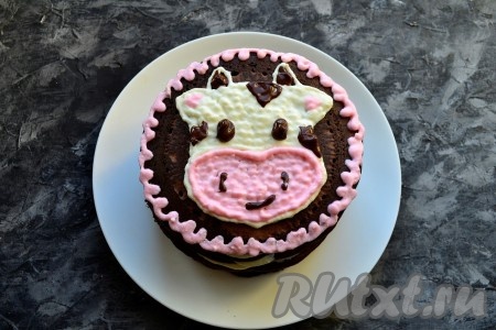 Затем маленькими точечками заполнить верхнюю часть головы бычка белым кремом, остальной крем покрасить в розовый цвет и также заполнить нижнюю часть головы, ушки и края тортика. Растопить шоколад, остудить и также из мешка с обрезанным кончиком нарисовать глазки, рожки, ротик и ноздри бычка.
