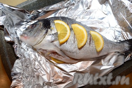 По прошествии времени возьмите достаточный кусок пищевой фольги (чтобы можно было в него завернуть рыбку целиком). Выложите фольгу в форму для запекания. Оставшуюся четверть лимона нарежьте на дольки. Половину долек выложите на фольгу. Поверх лимонных долек выложите замаринованную дораду и сверху разложите оставшиеся дольки лимона.
