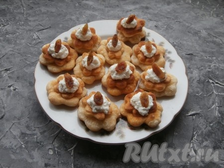 Если в твороге большие крупинки, можно всю массу немного измельчить погружным блендером. Творожную массу переложить в кондитерский мешок, срезать кончик и выдавить её в серединку пончиков на маленький цветок. На творожной начинке разместить по ягодке изюма.
