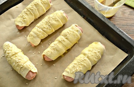 Яичный желток слегка взбить вилкой, а затем смазать им слоёное тесто. Присыпать, по желанию, сосиски в тесте кунжутом.