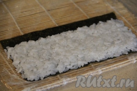 Я предварительно разрезаю лист нори на две части. На циновку (я оборачиваю циновку целлофаном) выложить одну половину листа нори блестящей стороной вниз. Часть остывшего риса равномерно распределить по нори, оставив, примерно, 1 см одного широкого края пустым Если рис сильно липнет к рукам, можете смочить руки немного водой.
