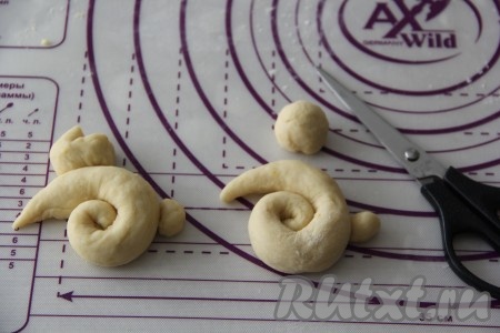 По прошествии 30 минут поднявшееся дрожжевое тесто ещё раз обмять и разделить на 7-8 частей (в результате получатся 7-8 пасхальных зайчиков). Приступаем к формированию первого "зайчонка", для этого нужно взять одну часть теста, разделить её на 3 части: большая часть - это будет тело зайчика, поменьше часть - это голова, самая маленькая часть - хвостик. Из большой части теста скрутить жгутик с тонкими кончиками, скрутить жгутик в улитку (как на фото). Сформировать колобок для головы, ножницами сделать разрез и у нас получатся ушки. Ну и хвостик сформировать в виде маленького колобка.