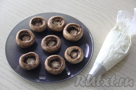 Переложить творожный сыр, смешанный с чесноком, в кулинарный мешок с фигурной насадкой. При желании можно обойтись без кулинарного мешка и выкладывать творожный сыр в шляпки шампиньонов ложкой.