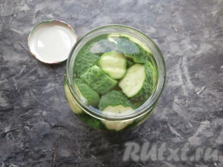 Залить огурцы кипятком, оставить, накрыв прокипячённой крышкой, на 10 минут. Затем воду слить в раковину, снова полностью залить нарезанные огурчики кипящей водой, оставить на 5 минут.