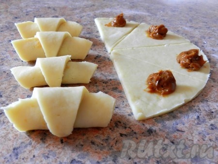 Ближе к основанию каждого сегмента, не забывая отступить от краёв, выложить по 1 чайной ложке варёной сгущёнки. Свернуть каждый сегмент, начиная со стороны с начинкой, в рулетик, формируя рогалик.