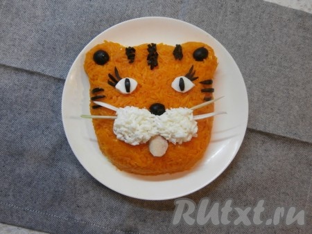 Осталось оформить только мордочку тигрёнка. Из части белка вырезать глаза, оставшиеся белки натереть на мелкой терке и выложить их в части носа (как на фото). Носик, зрачки, реснички, середину ушек и полоски сформировать с помощью нарезанных маслин. Язычок вырезать из ломтика колбасы. Усы сделать их белой части зелёного лука.