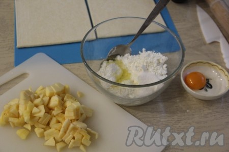 Предварительно разморозить слоёное тесто при комнатной температуре. Яблоки очистить от кожуры, удалить сердцевину, а затем нарезать на мелкие кубики. Сырое яйцо разделить на белок и желток. Соединить творог, сахар и белок. Желток нам потребуется позже для смазывания заготовок слоек.