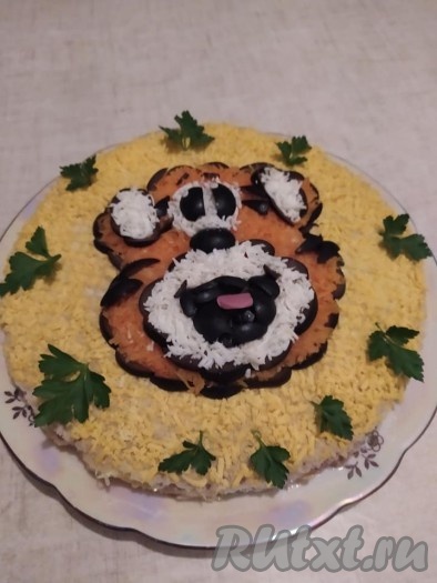Из маслин сделать контур для ушей и мордочки. Заполнить яичным белком. Глаза, нос и рот выложить из кусочков маслин, язык - кусочек колбаски. Украсить закусочный торт зеленью.