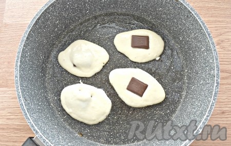 На сковороду вливаем, примерно, 2-3 столовые ложки масла и разогреваем его. Тесто столовой ложкой выкладываем на сковороду. В середину каждой заготовки оладушка кладём по шоколадной дольке. Сверху закрываем шоколад половиной столовой ложки теста.
