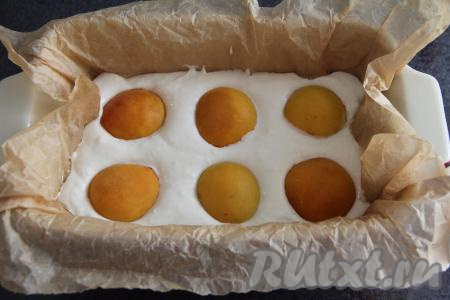 Форму для выпекания (я взяла прямоугольную форму размером 28 сантиметров на 15 сантиметров) застелить пергаментом и выложить творожную массу. Абрикосы разрезать на две части и удалить косточки. Выложить абрикосы поверх творожной массы срезом вниз и слегка их вдавить.