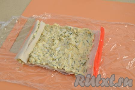 Таким образом внахлёст выложить 4 крабовых палочки. Если смотреть на фото, то красная часть у нас будет справа. Выложить 1/3 часть начинки из творожного сыра и яиц на крабовые палочки, пару сантиметров справа (там где красная часть палочки) оставить без начинки. Свернуть крабовые палочки в плотный рулет, начиная с левой стороны и помогая себе пищевой плёнкой.