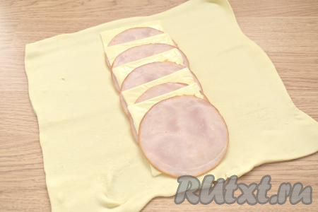 Подготовим нужные продукты для приготовления пирога с ветчиной и сыром.  Слоёное тесто (подойдёт как дрожжевое, так и бездрожжевое) достаём из морозилки и оставляем его размораживаться при комнатной температуре (мне на размораживание теста потребовалось чуть более 30 минут). Выкладываем размороженный пласт теста на стол, присыпанный мукой. Раскатываем тесто в квадратную заготовку толщиной 2-3 миллиметра. Сыр нарезаем на тонкие ломтики (я нарезала на 5 ломтиков). Отступая от верхней стороны квадрата 2-3 сантиметра, выкладываем по центру, чередуя, друг на друга ломтики ветчины и сыра.