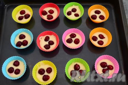 На противень для выпечки поставить силиконовые формы (так как формы у меня силиконовые, я их предварительно маслом не смазывала) и заполнить их на 1/3 объёма тестом (я выкладывала по 1 столовой ложке теста). В каждую форму для маффинов положить по 3 ягодки вишни, немного утопив их в тесто.