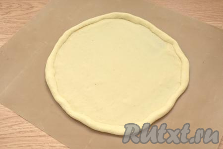 Поднявшееся тесто обминаем, делим на 2 равных части. Я сразу перекладываю 1 часть теста на бумагу для выпечки и раскатываю в круглый пласт. Края немного подгибаю к центру, как на фото. Получается заготовка для пиццы диаметром 24 сантиметра.