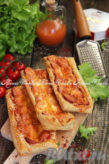 Закусочные слойки с начинкой из сосисок и сыра достать из духовки, переложить на тарелку, дать немного остыть и можно подавать к столу. Такая быстрая выпечка из слоёного теста станет и отличным дополнением к чаю, и сытным перекусом в течение дня.