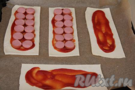Прямоугольные заготовки теста переложить на противень, застеленный ковриком (или пергаментом) для выпечки. Смазать середину каждой заготовки теста кетчупом, отступив от краёв по 1-2 сантиметра. Сосиски нарезать на кружочки толщиной 0,5 сантиметра и выложить на кетчуп. Смазать свободную часть теста взбитым желтком.