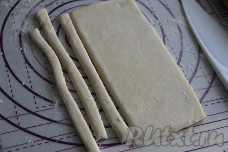 На поверхность, слегка присыпанную мукой, выложить разморозившееся слоёное тесто, слегка раскатать его скалкой, а затем нарезать на полоски шириной примерно 1 сантиметр.
