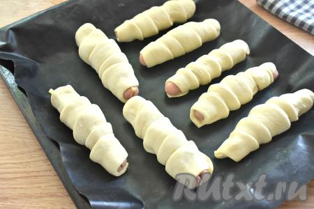 Оборачиваем полоску теста внахлёст вокруг сосиски с сыром, а затем выкладываем на противень, застеленный ковриком (или пергаментом) для выпечки. Постепенно таким образом формируем все сосиски в тесте. 