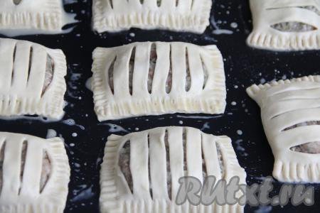 Тщательно скрепить края каждого пирожка с помощью вилки (если вилка будет сильно липнуть к слоёному тесту, тогда слегка обмакните её в муку). Переложить пирожки с фаршем на противень, заранее застеленный ковриком (или пергаментом) для выпечки. Яичный желток в небольшой ёмкости слегка взбить вилкой. Сверху смазать пирожки взбитым желтком.