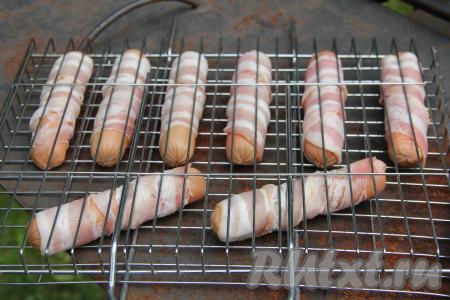  Заранее подготовить мангал, угли должны быть хорошо прогревшимися, но без прямого огня. Выложить сосиски в беконе на решётку. Поместить решётку на мангал с горячими углями.